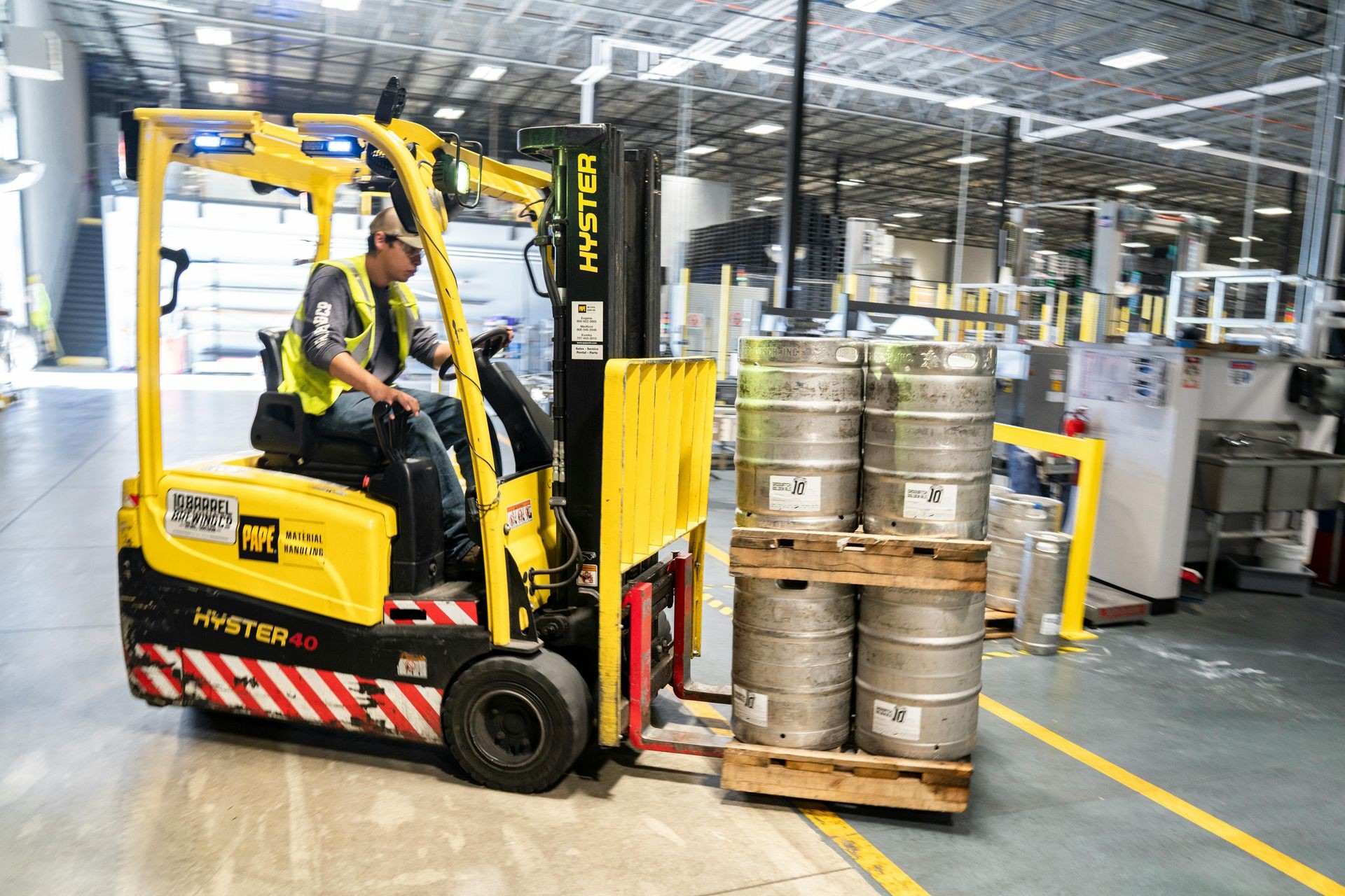 Worker operating a yellow forklift carrying metal kegs in a spacious industrial warehouse.